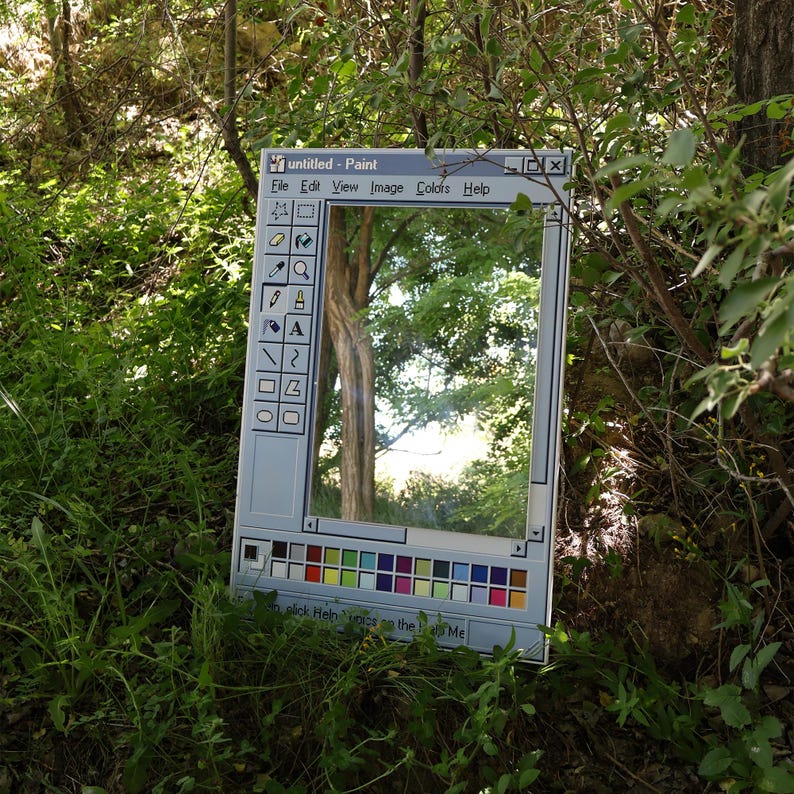 以下が含まれることがあります： クラシックなWindowsペイントアプリケーションのインターフェースに似せてフレームされた長方形の鏡。鏡は、木や葉のある自然な屋外のシーンを映し出しています。フレームは、カラーパレットとツールアイコンを備えたライトグレーです。