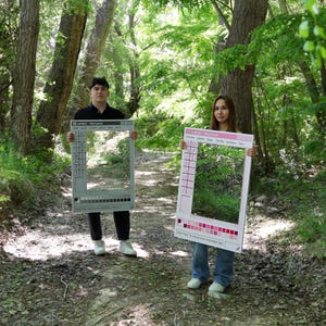 May include: Two people holding large, rectangular photo booth props in a forest setting. One prop is gray with a computer interface design, the other is pink with a color bar and the text "Dreams Sparkle Magic Twinkle Rainbow Fwy".