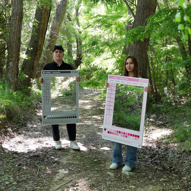 May include: Two people holding large, rectangular photo booth props in a forest setting. One prop is gray with a computer interface design, the other is pink with a color bar and the text "Dreams Sparkle Magic Twinkle Rainbow Fwy".