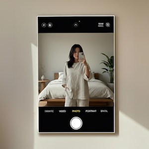 May include: A framed image of a person taking a selfie with a smartphone. The person is wearing a light-colored linen outfit. The phone's camera interface is visible, with options for cinematic, video, photo, portrait, and spatial modes.