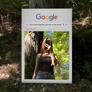 以下が含まれることがあります： 「The most beautiful woman in the world」という検索クエリが表示されたGoogle検索バーのあるフレーム画像。フレーム内の画像は、黒いトップスとジーンズを着た人が顔の前に携帯電話を持っている様子を示しています。