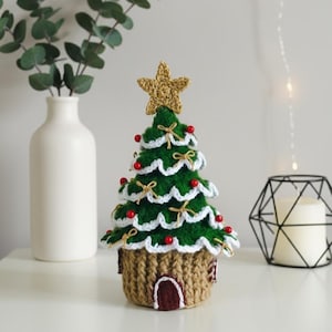 Puede incluir: Decoración de árbol de Navidad de ganchillo hecha a mano. El árbol es verde con ribete blanco, adornos de cuentas rojas y una estrella dorada. La base es una casa marrón con una puerta marrón. Un acento festivo navideño.