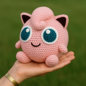 May include: A handmade, pink crochet Jigglypuff plushie with large blue and white eyes, small arms, and rounded feet. The plushie has black ear tips and a curled tuft of hair on top. The toy is held in a hand against a blurred green background.