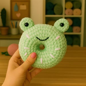 May include: A light green crocheted frog-shaped donut. The donut features a central hole, pink sprinkles, and black eyes. The frog donut is held in a hand.