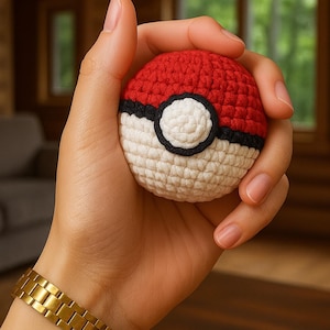 Könnte beinhalten: Ein gehäkelter Pokéball in Rot, Weiß und Schwarz. Der Ball wird in einer Hand gehalten, was die detaillierten Stiche und das Design zeigt. Der Pokéball hat eine weiße Mitte mit einem schwarzen Rand und einer roten Oberseite. Das Thema bezieht sich auf die beliebte Anime-Serie.