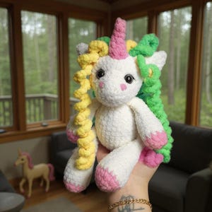 Può includere: Un unicorno bianco fatto a mano all'uncinetto con un corno rosa, zoccoli rosa e capelli ricci colorati in giallo e verde. L'unicorno ha occhi neri e guance rosa. Sullo sfondo è visibile una statuina di unicorno più piccola.