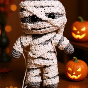 May include: A handmade crochet mummy doll, with black button eyes, wrapped in white and grey yarn. Two carved orange pumpkins with lit faces sit nearby, suggesting a Halloween theme. The doll is approximately 15 cm tall.