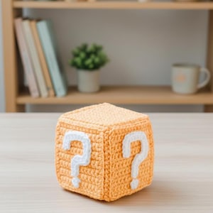 May include: A light orange crocheted cube featuring white question marks on two sides. The handmade cube is placed on a light-coloured wooden surface. The cube is a decorative item, possibly a toy or a gift.