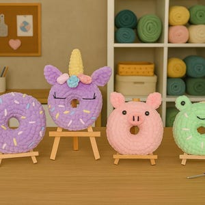 May include: Four crocheted donut-shaped toys. A purple donut, a unicorn donut, a pink pig donut, and a green frog donut. Each donut has a different design and is displayed on a small wooden easel.