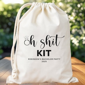 May include: A cream-colored drawstring bag with the text "Oh shit KIT" in black script and block letters. The bag includes the text "ROBINSON'S BACHELOR PARTY 2025". The bag is sitting on a wooden table.