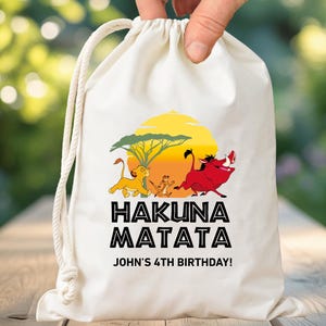 May include: A white drawstring bag featuring a colorful graphic of characters from The Lion King, with the words "HAKUNA MATATA" and "JOHN'S 4TH BIRTHDAY!" printed in black. The bag is held up by a hand.