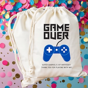 May include: Two cream-coloured drawstring bags with the words "GAME OVER" in black block letters and a blue video game controller graphic. Below the graphic is the text "SUPER GABRIEL'S 1ST BIRTHDAY! THANK YOU FOR PLAYING WITH ME!"