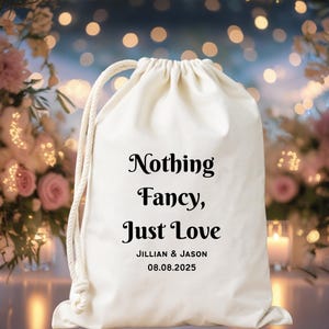 May include: Cream-colored drawstring bag with the text "Nothing Fancy, Just Love" in elegant black script. The bag includes the names "JILLIAN & JASON" and the date "08.08.2025". The background features soft floral arrangements.