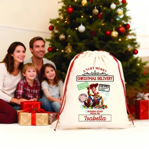 以下が含まれることがあります： 赤色の紐とトリムが付いた、オフホワイトの大きなクリスマスギフトバッグ。「A Very Merry Christmas Delivery」の文字と、トイ・ストーリーのウッディの漫画の画像が特徴です。「Isabella」という名前が下に印刷されています。