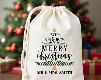 Puede incluir: Una bolsa de regalo crema con cordón con el texto "We wish you a Merry Christmas" en escritura negra. El diseño incluye una forma de árbol de Navidad, una estrella dorada y los nombres "MR & MRS. SMITH" en la parte inferior. Un árbol de Navidad está en el fondo.