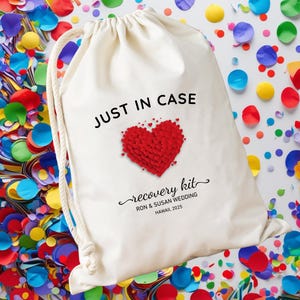 May include: A cream-colored drawstring bag with the text "JUST IN CASE" in black. Below is a red heart made of smaller hearts, and the words "recovery kit" in script. The bag is for the Ron & Susan wedding in Hawaii, 2025.