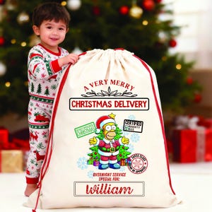 May include: A child in festive pyjamas stands beside a large cream Christmas gift bag with red trim. The bag displays the text "A Very Merry Christmas Delivery" and a cartoon character dressed as Santa Claus. The name "William" is printed on the bag.