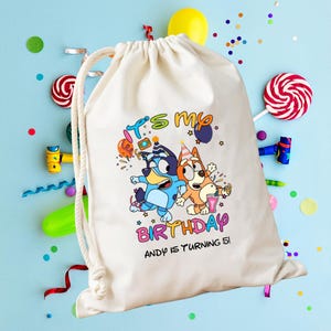 Peut inclure: Un sac à cordon blanc avec un graphisme coloré indiquant "C'est mon anniversaire" avec deux chiens de dessin animé. Le texte "Andy a 5 ans !" est en dessous. Le sac est sur une surface bleu clair avec des décorations de fête.