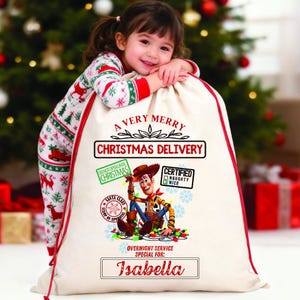 以下が含まれることがあります： 赤色の縁取りと巾着開閉部が付いた、オフホワイトの大きなクリスマスギフトバッグ。「A Very Merry Christmas Delivery」の文字と、トイ・ストーリーのウッディの漫画の画像が特徴です。「Isabella」という名前が下に印刷されています。