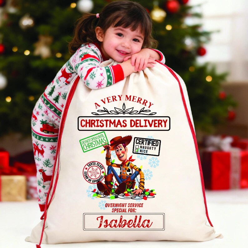 以下が含まれることがあります： 赤色の縁取りと巾着開閉部が付いた、オフホワイトの大きなクリスマスギフトバッグ。「A Very Merry Christmas Delivery」の文字と、トイ・ストーリーのウッディの漫画の画像が特徴です。「Isabella」という名前が下に印刷されています。