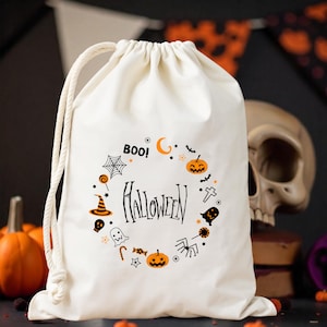 Puede incluir: Bolsa con cordón de color crema con un diseño de Halloween. El diseño incluye la palabra "Halloween" rodeada de calabazas naranjas, murciélagos, fantasmas y otros elementos espeluznantes. También se incluye la palabra "Boo!".