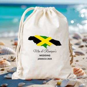 May include: A cream-colored drawstring bag featuring the Jamaican flag in black, yellow, and green. The bag is printed with the text "Mia & Ranique's WEDDING JAMAICA 2025". The bag is on a sandy beach with seashells.