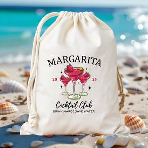 May include: A cream-coloured drawstring bag featuring the word "MARGARITA" in black script. Below, a graphic of three margarita glasses with strawberries and the text "Cocktail Club" and "DRINK MARGS SAVE WATER". The bag has a white drawstring.
