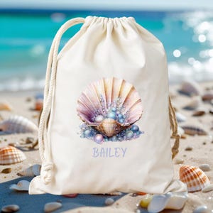 May include: A cream-colored drawstring bag featuring a watercolor seashell design with pearls in shades of pink, purple, and blue. The name "BAILEY" is printed in a light purple script below the seashell. The bag is on a sandy beach.