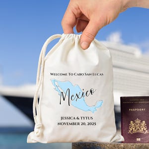 May include: A cream drawstring bag featuring the words "WELCOME TO CABO SAN LUCAS" and "Mexico" in black script, with a light blue map of Mexico. The bag also includes the names "JESSICA & TYTUS" and the date "NOVEMBER 20, 2025".