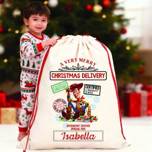 以下が含まれることがあります： 赤色の縁取りと巾着が付いた、オフホワイトの大きなクリスマスギフトバッグ。「A Very Merry Christmas Delivery」の文字と、トイ・ストーリーのウッディのグラフィックが特徴です。「Isabella」という名前が下部に印刷されています。