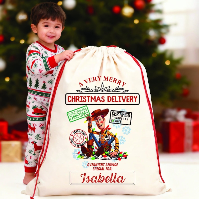 以下が含まれることがあります： 赤色の縁取りと巾着が付いた、オフホワイトの大きなクリスマスギフトバッグ。「A Very Merry Christmas Delivery」の文字と、トイ・ストーリーのウッディのグラフィックが特徴です。「Isabella」という名前が下部に印刷されています。