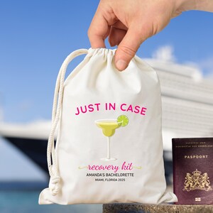 May include: A cream drawstring bag with the text "JUST IN CASE" and "recovery kit" printed in pink and yellow. The bag features a margarita illustration and the text "AMANDA'S BACHELORETTE MIAMI, FLORIDA 2025". A passport is visible next to the bag.