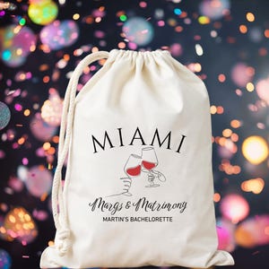 May include: A cream-coloured drawstring bag with the word "MIAMI" in black script. Below, it reads "Margs & Matrimony" and "MARTIN'S BACHELORETTE". The bag features a line drawing of two wine glasses with red wine, clinking together.