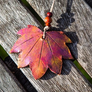 May include: A maple leaf pendant with a vibrant color scheme of red, orange, and purple. The leaf is suspended by a beige cord with wooden beads. The pendant is resting on a weathered wooden surface.