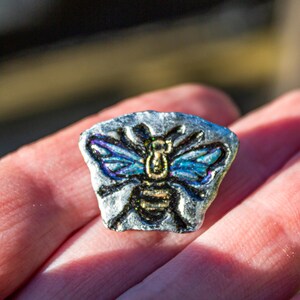 May include: A small, hand-painted, bee-shaped pendant. The bee has blue and black wings, a gold body, and is set against a silver-colored background. The pendant is held in a person's hand.