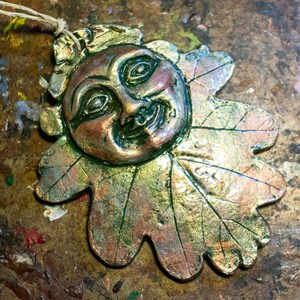 May include: A ceramic ornament shaped like an oak leaf with a smiling face in the center. The leaf is painted in shades of green, gold, and brown.
