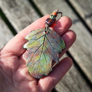 May include: A handcrafted leaf pendant with a detailed vein pattern. The pendant features a blend of green, bronze, and copper tones. It is suspended from a cord with decorative beads. The leaf measures approximately 2.5 inches in length.