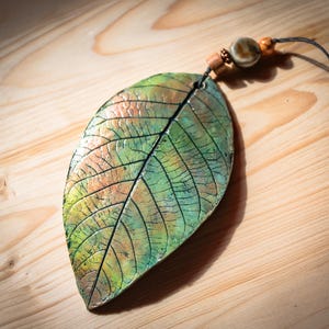 May include: A handcrafted leaf pendant with a naturalistic design. The pendant features a detailed leaf shape with green, orange, and brown hues, accented by black veins. It hangs from a black cord with wooden and ceramic beads.