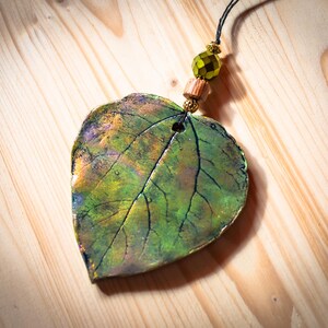 May include: A handcrafted leaf pendant with a naturalistic design. The pendant is primarily green with hints of yellow and purple, featuring detailed veining. It hangs from a black cord with decorative beads, including a faceted green bead and wooden accent.