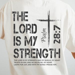 May include: Cream-colored t-shirt with the text "THE LORD IS MY STRENGTH" in black, along with a cross graphic and the Bible verse Psalm 28:7. The shirt also includes a quote from the verse.