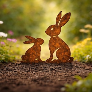 Könnte beinhalten: Zwei rostige Metall-Kaninchen-Silhouetten, eine große und eine kleinere, sitzen auf einem Beet aus dunkelbrauner Erde. Die Kaninchen befinden sich in einem Garten mit verschwommenem grünen Laub und bunten Blumen im Hintergrund.