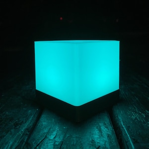 May include: A square, glowing teal light with a black base. The light is cube-shaped and emits a soft, even glow. The object is sitting on a dark, textured surface.