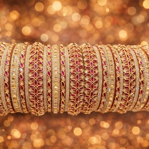 Könnte beinhalten: Eine Sammlung goldener Armreifen mit aufwendigen Designs. Jeder Armreif weist ein Muster aus roten Edelsteinen und klaren Kristallen auf, die auf einem goldenen Grund gesetzt sind. Die Armreifen werden auf einem Holzstab vor einem verschwommenen, goldenen Hintergrund präsentiert.