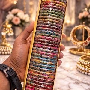 May include: A selection of vibrant bangles in a yellow box. The bangles feature detailed patterns and a spectrum of colours, including pink, blue, green, and gold. The box is held in a hand, with other jewellery visible in the background.