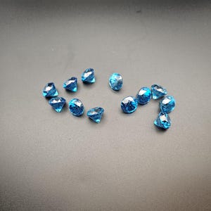 Puede incluir: Una colección de pequeños diamantes azules facetados de acrílico dispersos sobre una superficie gris oscuro. Las gemas varían en forma, algunas parecen más puntiagudas y otras más redondeadas. El efecto general es de brillo disperso y reflexión de la luz.