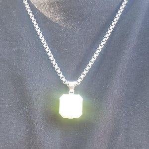 May include: Silver-toned necklace with a square pendant. The pendant has a bright, reflective surface and is suspended from a chain with a box link design. The necklace is displayed against a dark background.