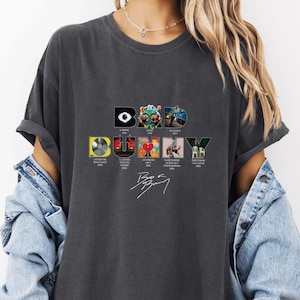 May include: Dark gray t-shirt featuring the text "BAD BUNNY" in a stylized font, with album art graphics. The shirt includes the artist's signature. The design incorporates album covers and dates.