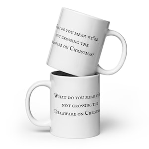May include: Two white ceramic mugs, one stacked on top of the other. The top mug is angled, and both mugs feature the text "WHAT DO YOU MEAN WE'RE NOT CROSSING THE DELAWARE ON CHRISTMAS?" printed in black.