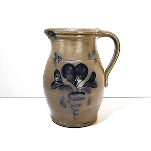 May include: A tan stoneware pitcher with a speckled texture and a single handle. The front of the vessel features a hand-painted cobalt blue floral design with a central heart-shaped motif and a spiral base, finished with a smooth glazed rim.