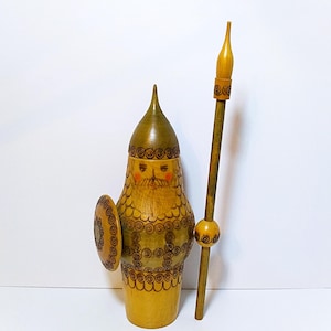 May include: A wooden figurine of a warrior wearing a pointed helmet, featuring a painted face with a beard, a circular shield attached to the side, and a long spear held upright. The surface is decorated with dark brown spiral patterns.